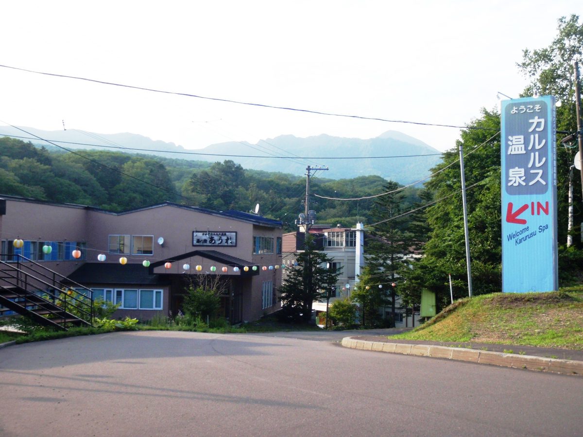 A welcome sign for Karurusu Onsen beside a road, with an inn building and forested mountains behind