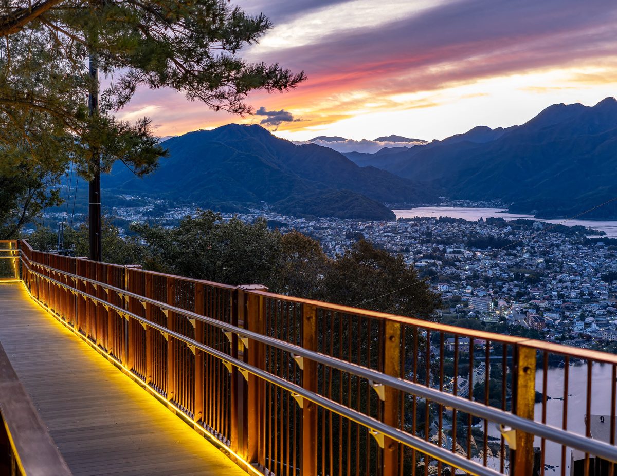 A lit wooden walkway with a railing overlooking a town and lake at dusk