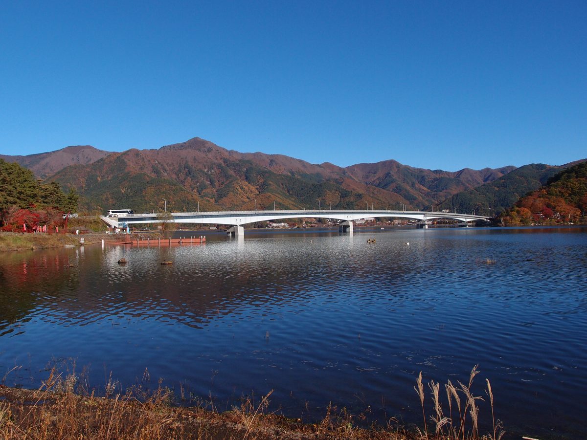 The Kawaguchiko Bridge crossing Lake Kawaguchi under a clear blue sky, with autumn-colored mountains behind