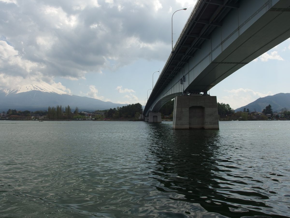 The underside of a long road bridge over Lake Kawaguchi, seen from the water, with mountains on the far shore