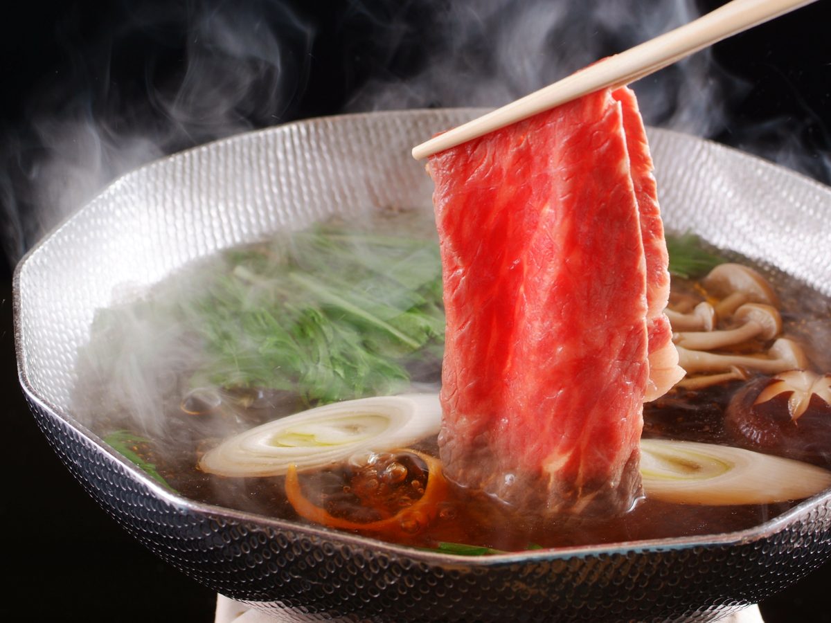 A slice of raw beef held with chopsticks over a steaming shabu-shabu pot