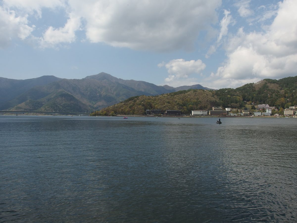 Lake Kawaguchi on a partly cloudy day, with wooded hills, a long bridge and small boats