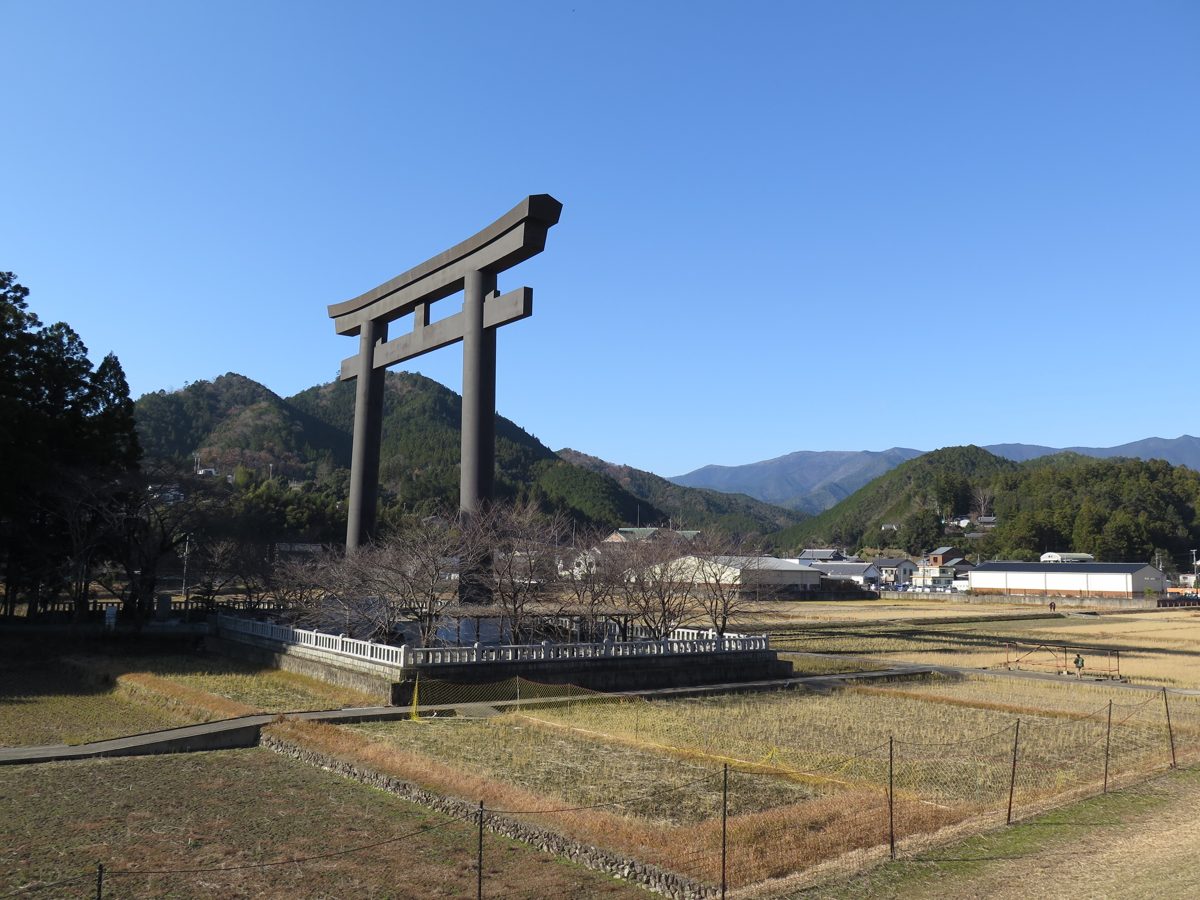 A giant torii gate standing in an open field under a blue sky, with wooded mountains behind