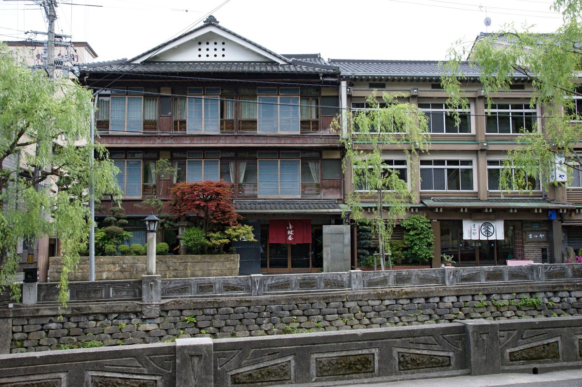Multi-story wooden inn buildings behind willow trees and a stone river wall in Kinosaki Onsen