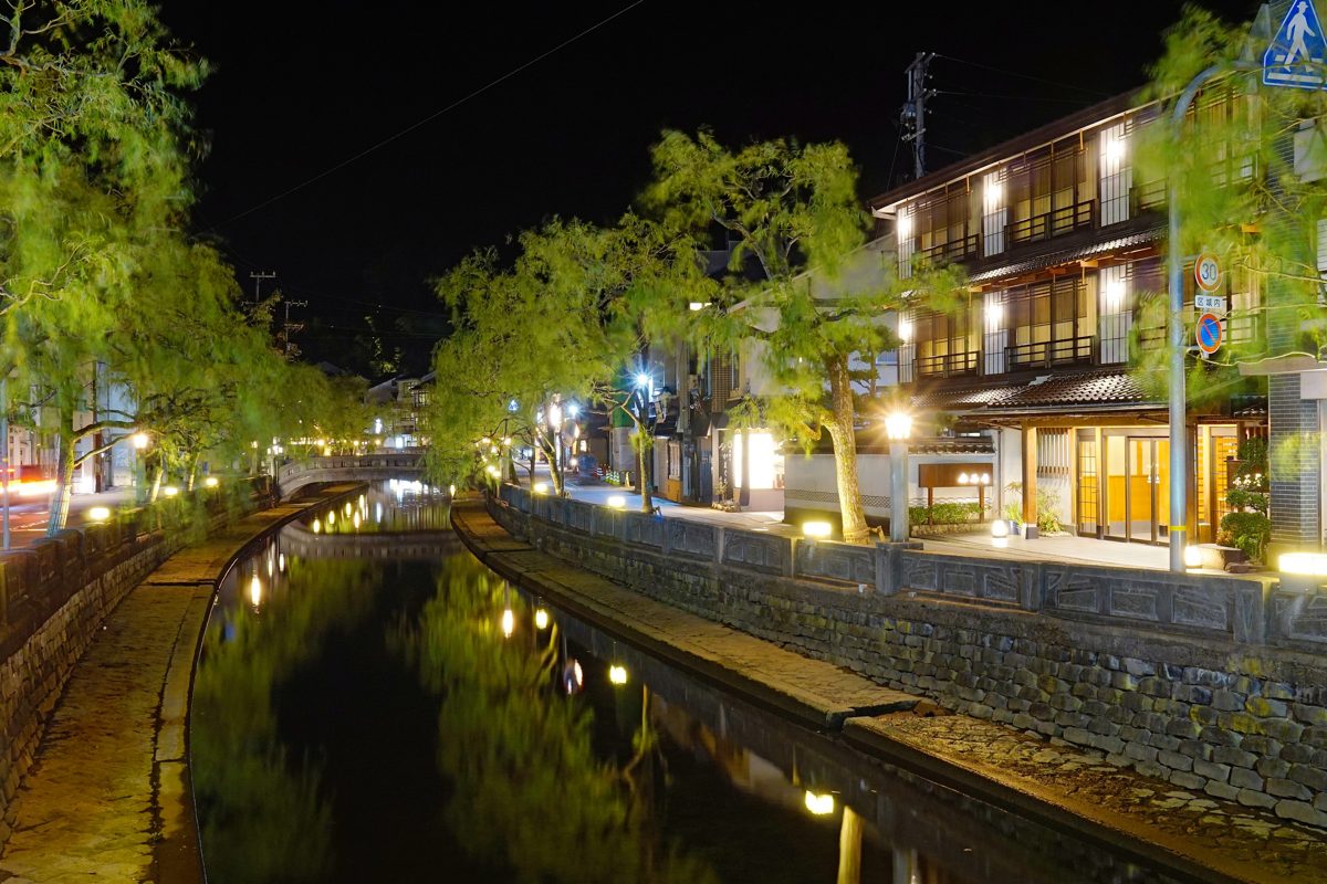 Lit-up willow trees and wooden buildings along the river in Kinosaki Onsen at night