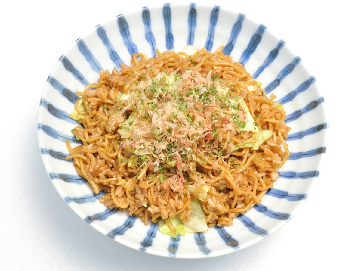 A striped bowl of brown fried noodles and rice topped with bonito flakes