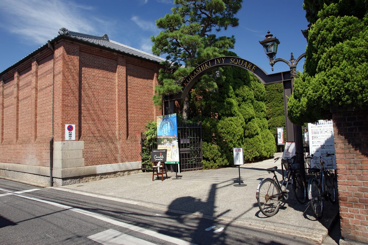 A red-brick building beside a black iron arch reading Kurashiki Ivy Square over an entrance gate, with bicycles parked nearby