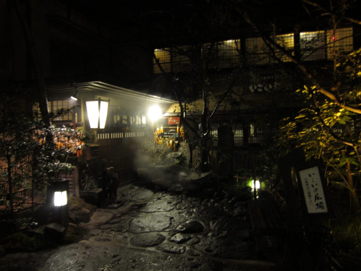 A lantern-lit stone path leading to a wooden building at night, with steam rising