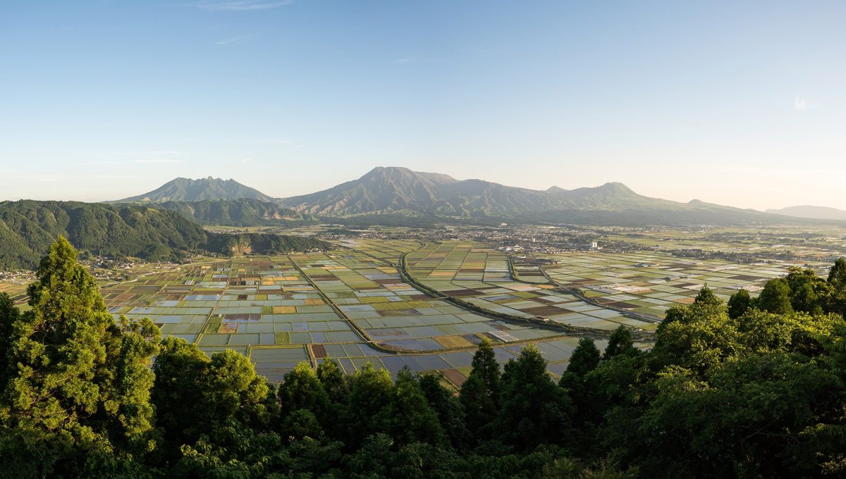 Wide daytime view over the Aso caldera with farmland, forest and mountains under a blue sky
