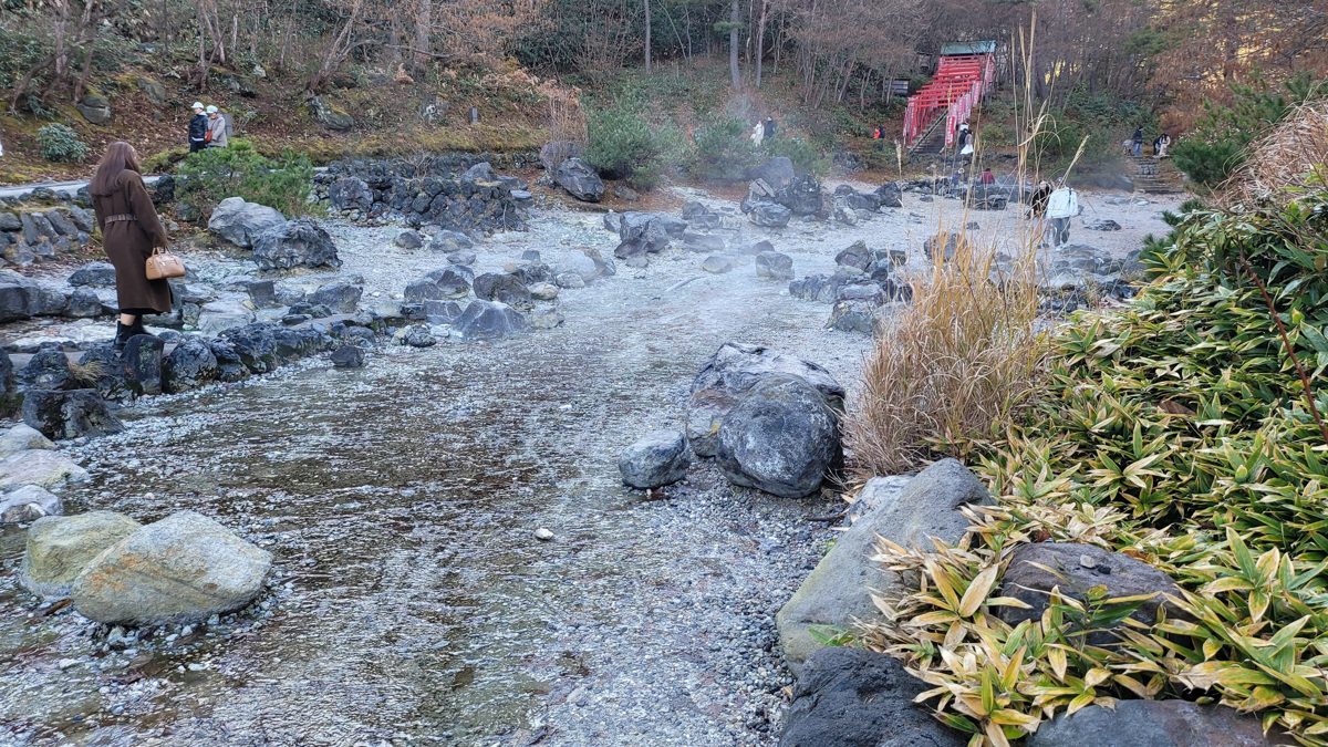 Steaming hot-spring water flowing over stones at Sainokawara Park in Kusatsu Onsen, with red torii gates on the wooded slope