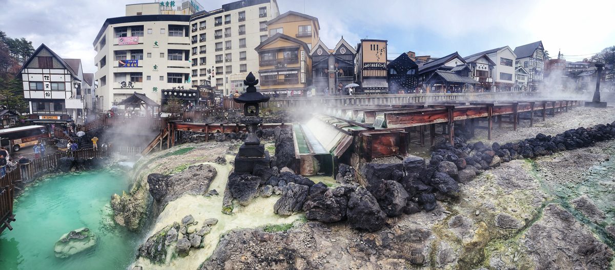 Turquoise hot water pooling among rocks at Yubatake with steam and town buildings behind