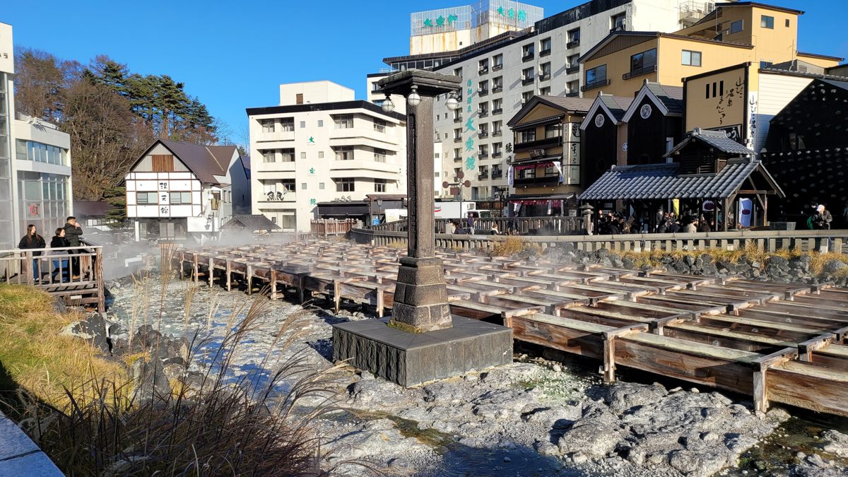 Wooden channels of Yubatake with hotels and shops around it under a blue sky