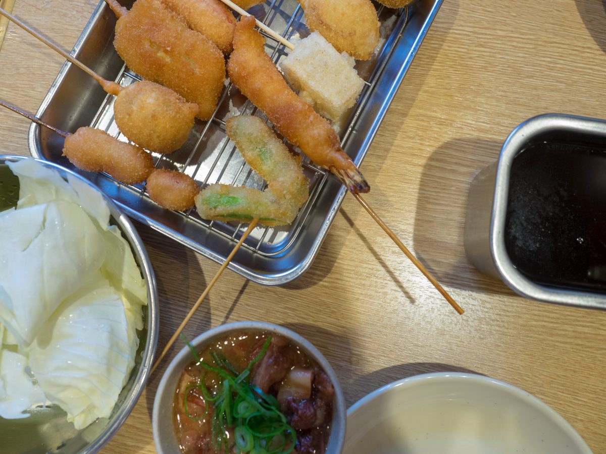 Deep-fried skewers on a wire rack with raw cabbage, a stew and a tin of dipping sauce