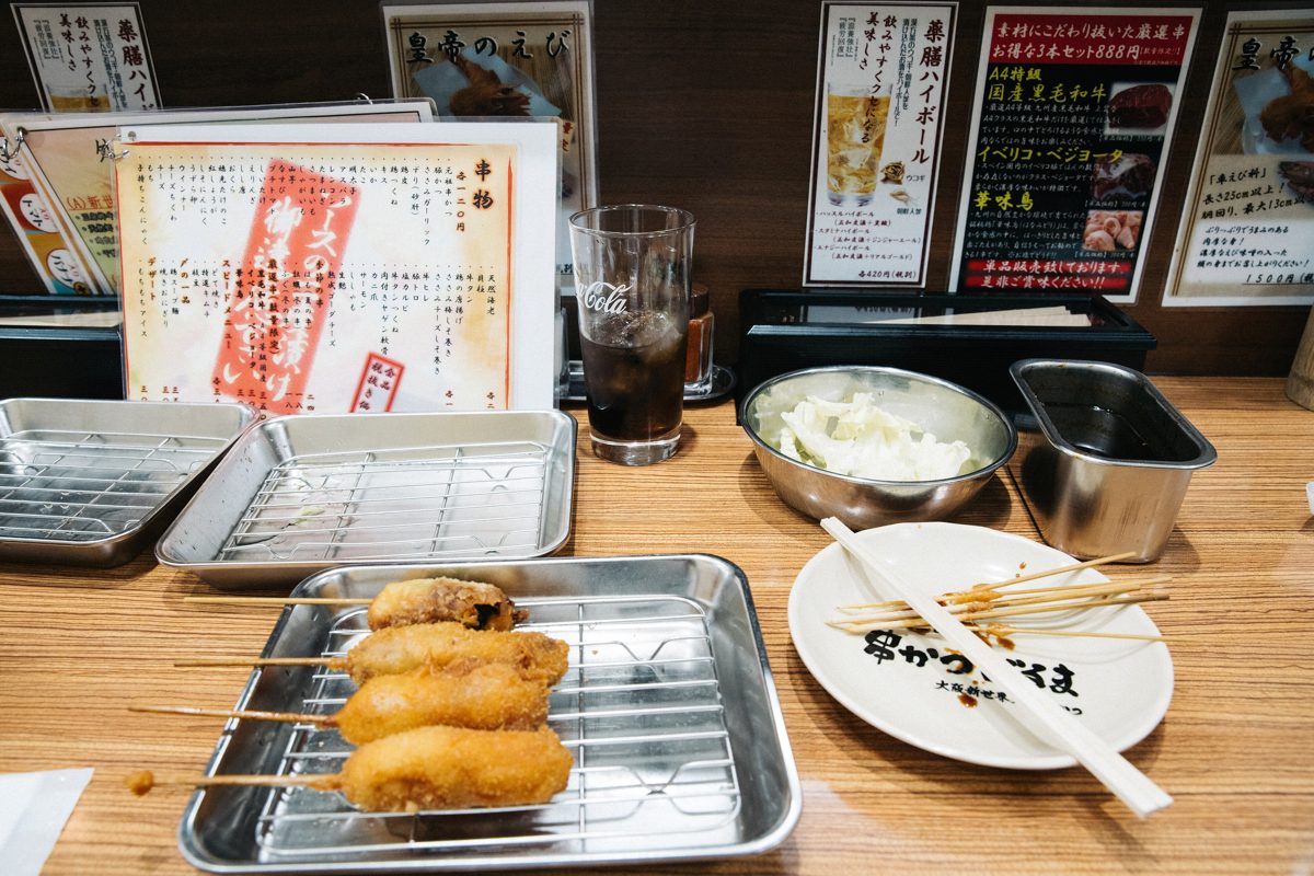 Kushikatsu skewers on a wire tray beside a metal sauce tin and a bowl of cabbage at a counter
