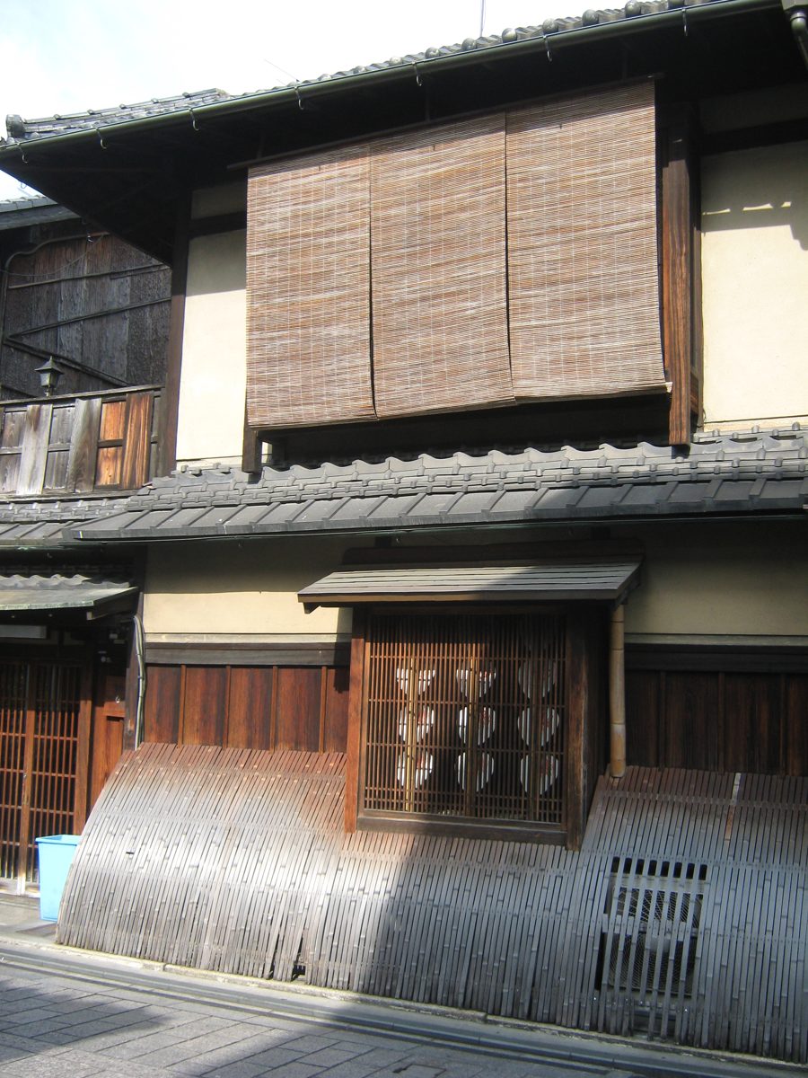A two-story wooden townhouse with a reed blind, a latticed window and a curved bamboo fence
