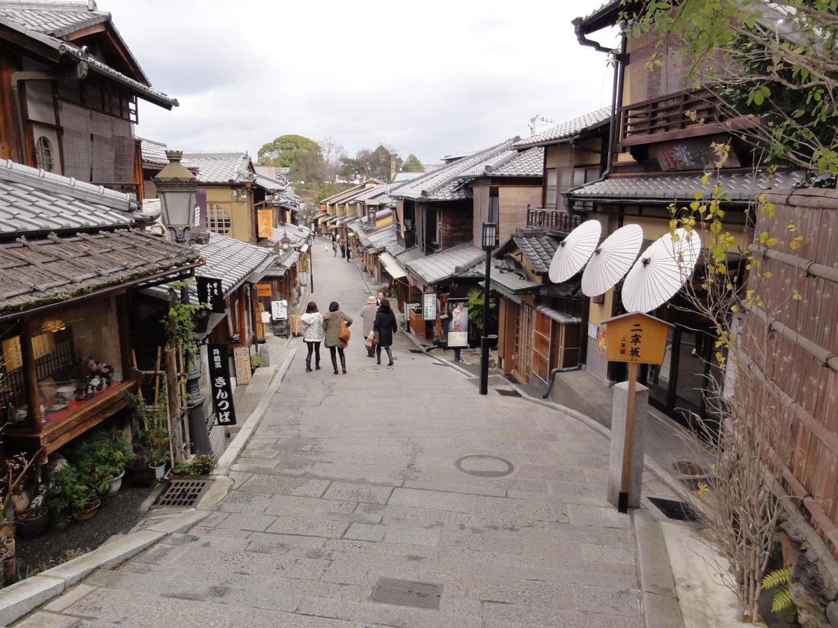A stone-paved lane lined with old wooden houses and shops in Kyoto, with a few people walking