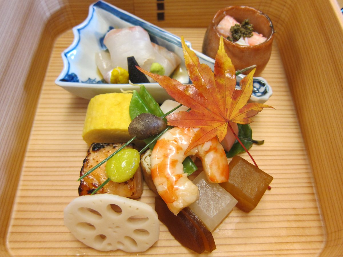 Small seasonal dishes arranged on a tray, including rolled omelet, shrimp, lotus root and a maple leaf