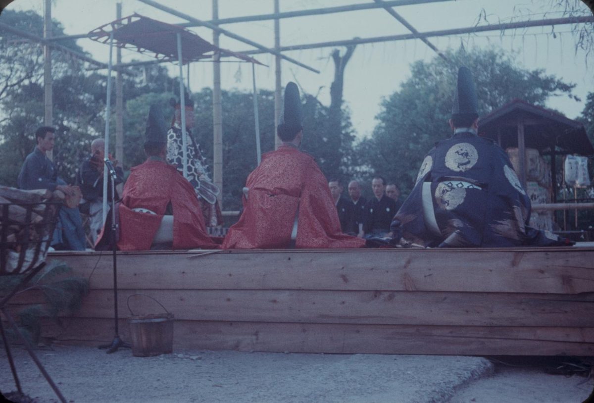 Noh performers in costume on an outdoor wooden stage, with musicians seated at the back