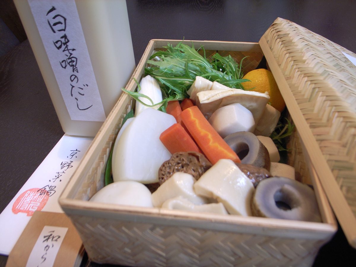 A bamboo basket of vegetables and other hot pot ingredients next to a bottle of white miso broth