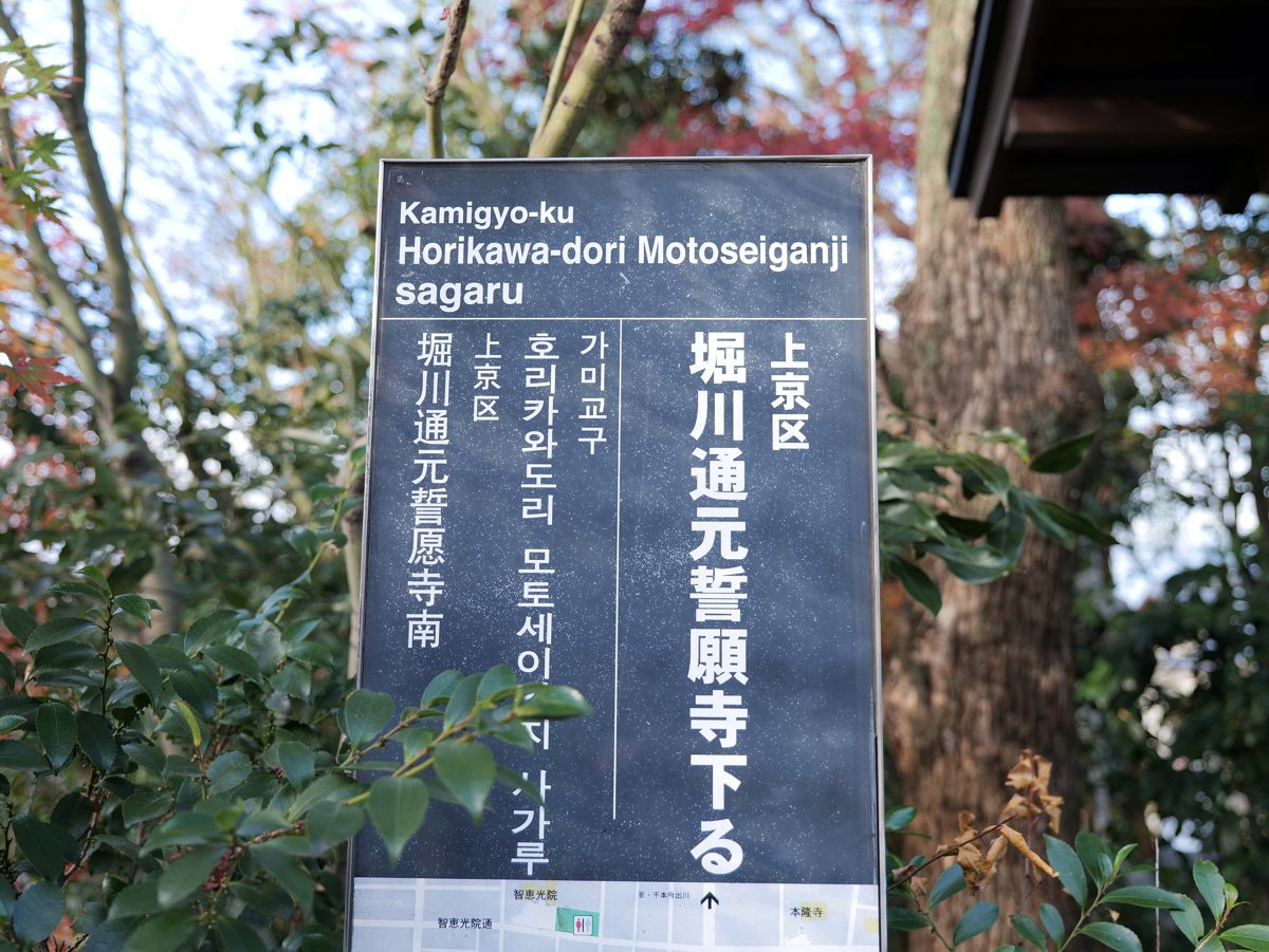 A dark blue street-address sign reading Kamigyo-ku Horikawa-dori Motoseiganji sagaru, with trees behind it