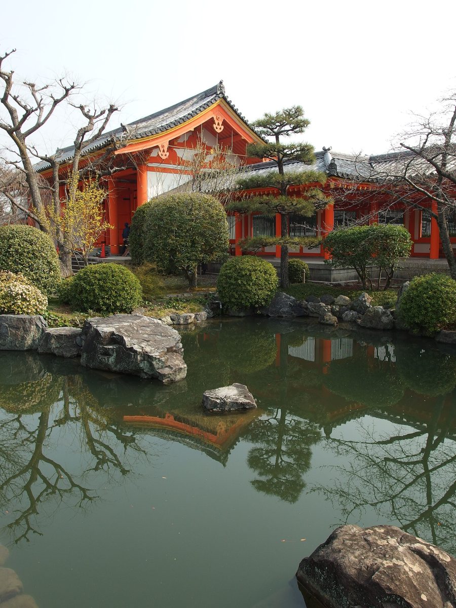 A pond with rocks and trimmed shrubs in front of red-painted temple buildings