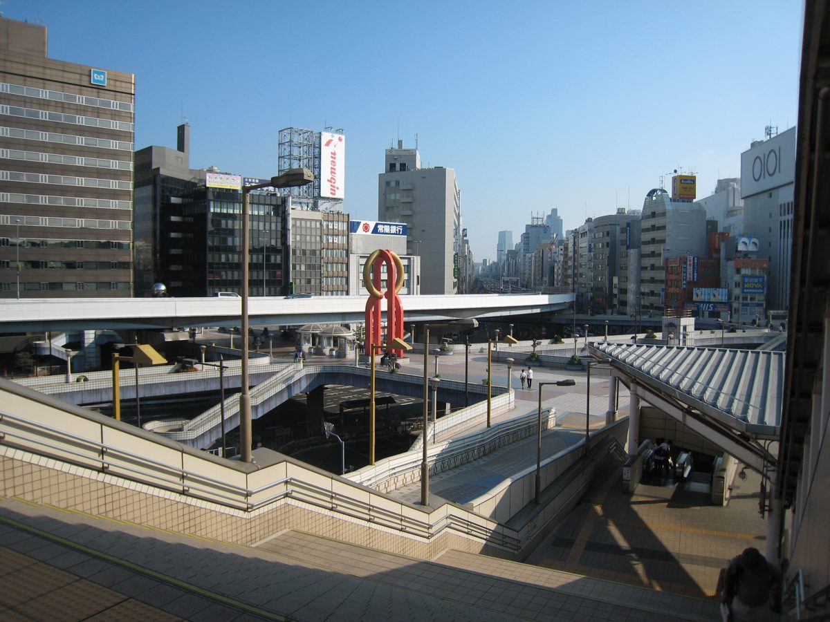A wide pedestrian deck beside a railway station with city buildings under a clear sky