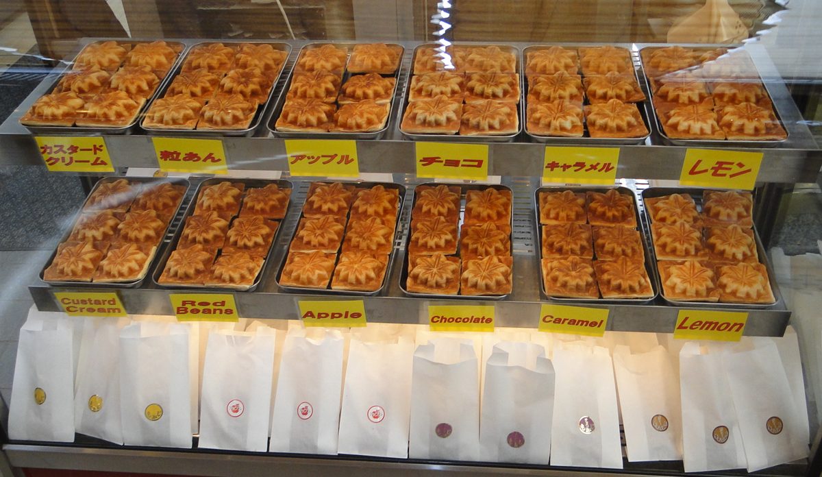 Trays of maple-leaf-shaped cakes in a shop case, labeled custard cream, red beans, apple, chocolate, caramel and lemon