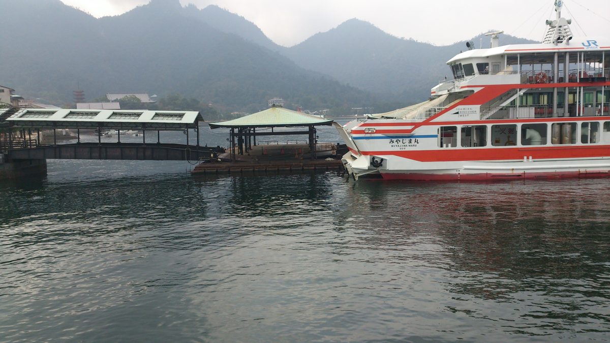 A red and white ferry beside the pier at the Miyajima ferry terminal, with hills behind