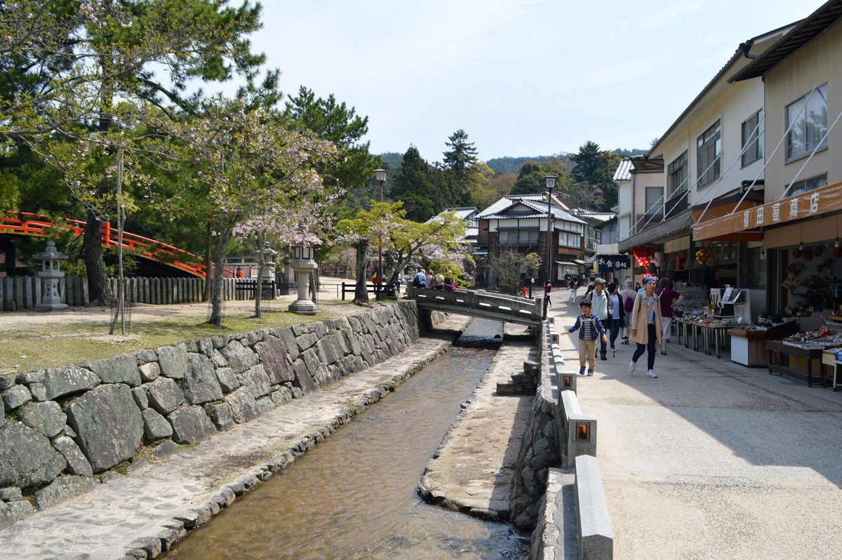 A stone-lined brook beside a street of shops on Miyajima on a sunny spring day, with an orange arched bridge on the left