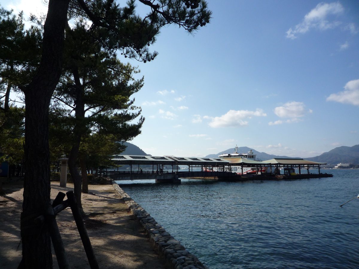 Pine trees on a seaside path beside a covered ferry pier with a docked ferry, mountains and blue sky behind