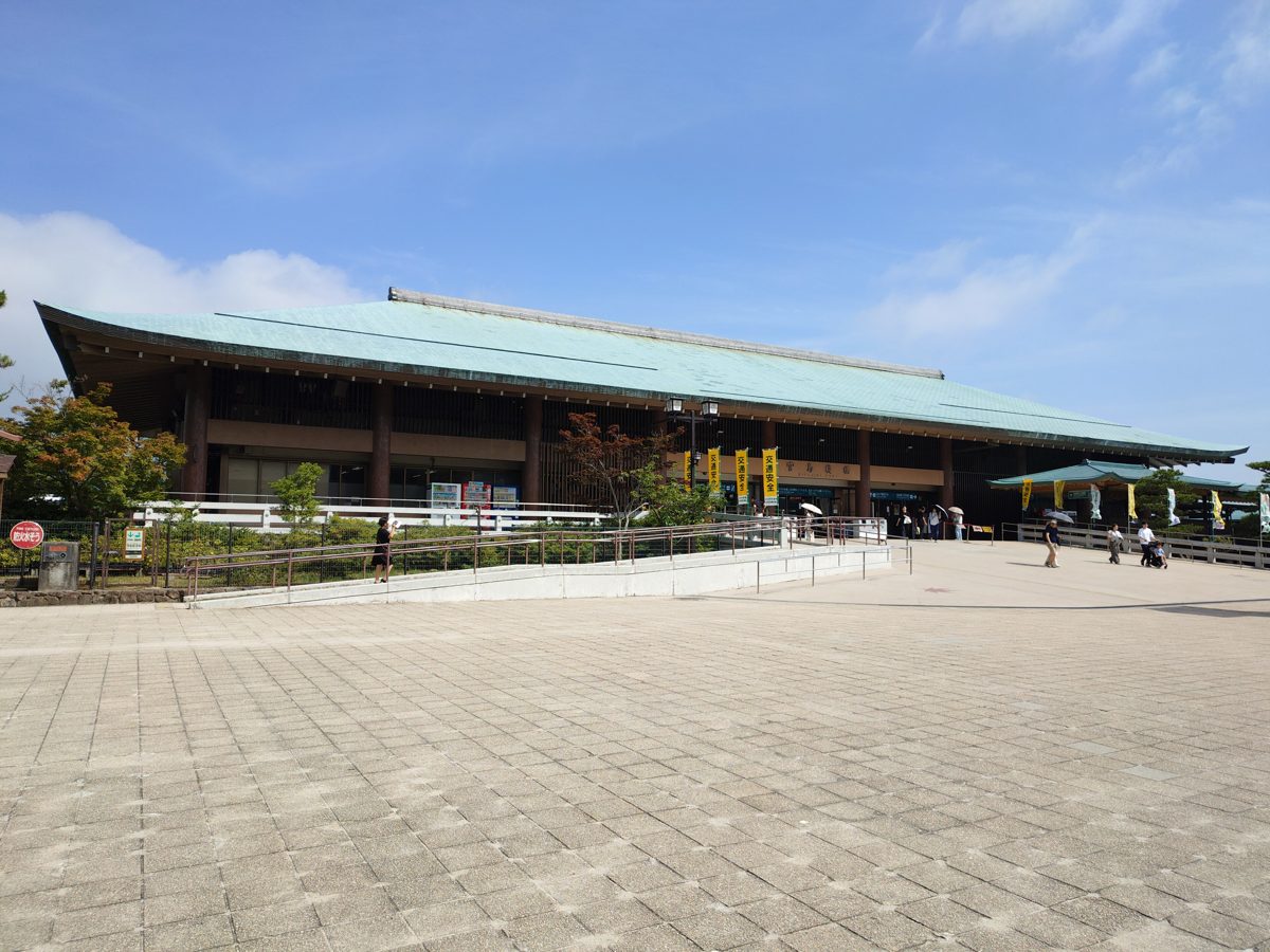 A large ferry terminal building with a pale green roof and a paved plaza under a blue sky on Miyajima