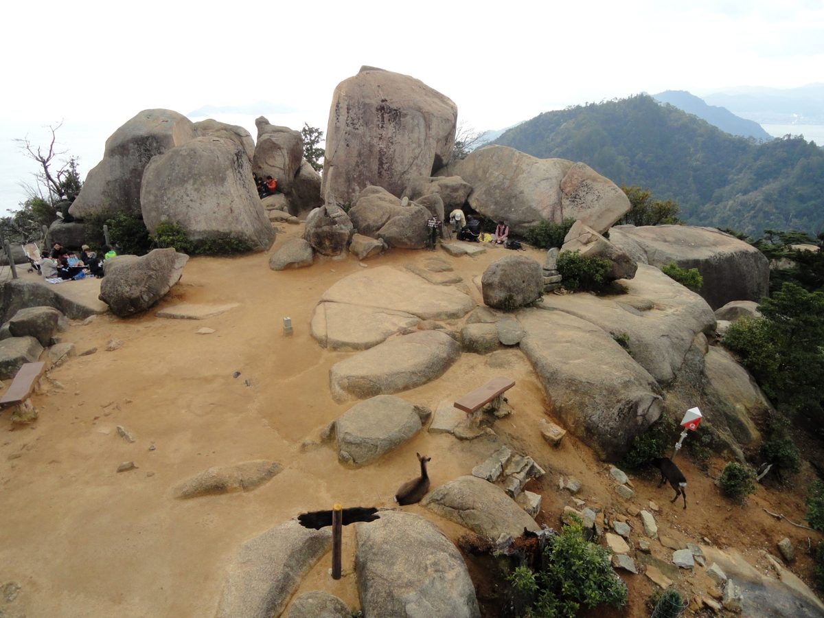 Large granite boulders and a sandy clearing at the summit of Mount Misen, with green hills and the sea behind