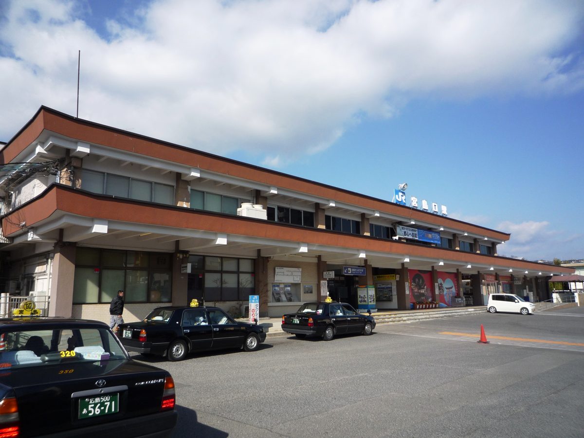 The two-story building of JR Miyajimaguchi Station with taxis parked in front under a blue sky