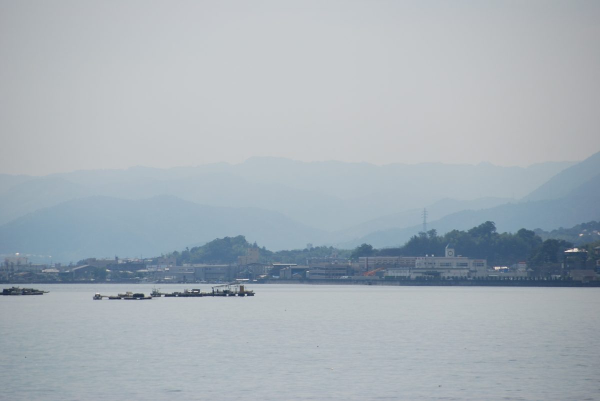 Calm gray sea with oyster rafts and a low coastline under hazy mountains