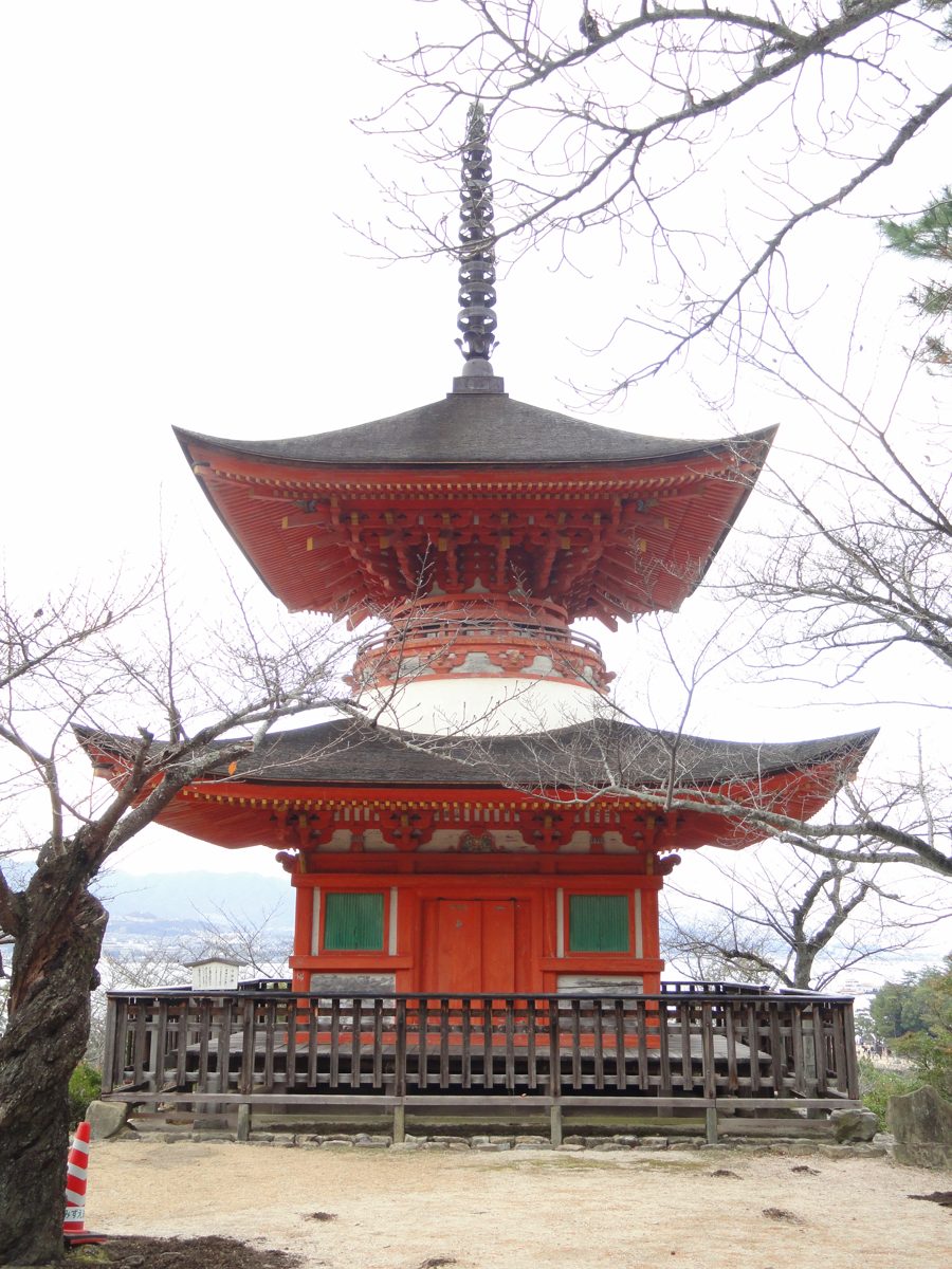 A two-tiered orange pagoda with a tall spire behind a wooden fence and bare branches