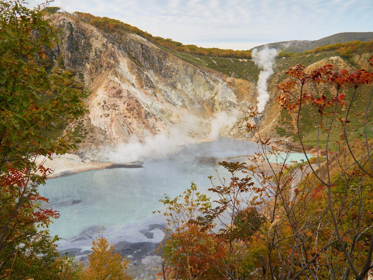 A steaming gray-blue pond below a pale crater wall with autumn leaves in the foreground