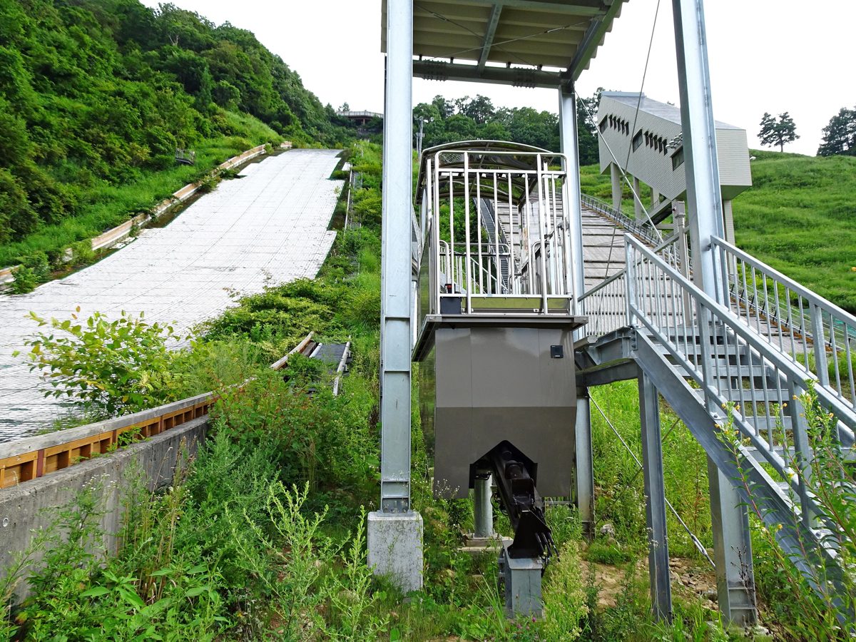 A slope car rail and stairs beside a ski jump hill covered in green grass in summer