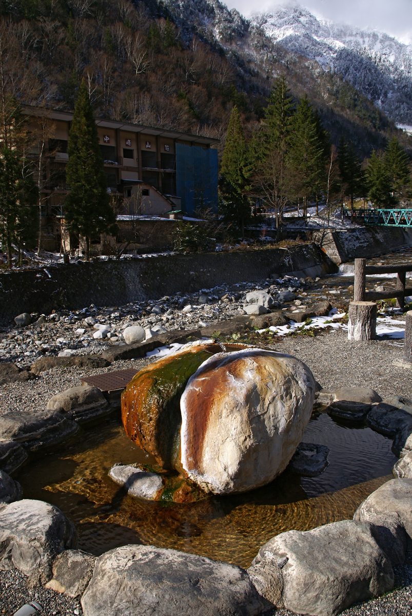 Hot spring water running over a stained rock in a stone pool, with a river and snowy mountains behind