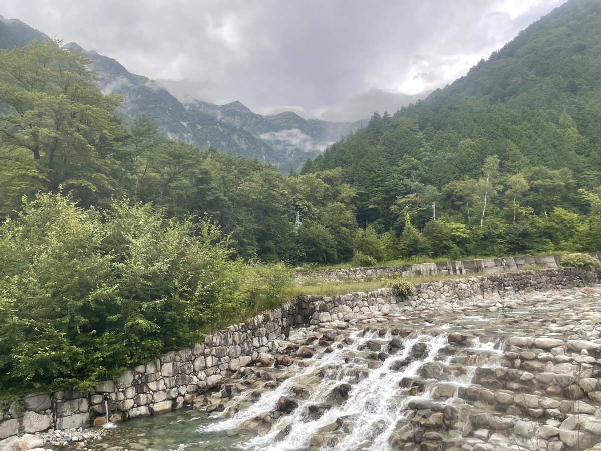 A rocky river with small cascades between forested mountains under clouds