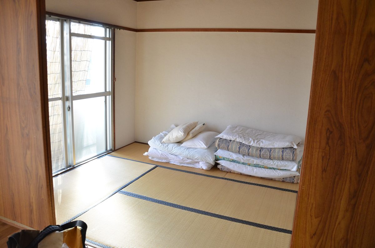 A plain tatami room with folded futon bedding and pillows stacked against the wall