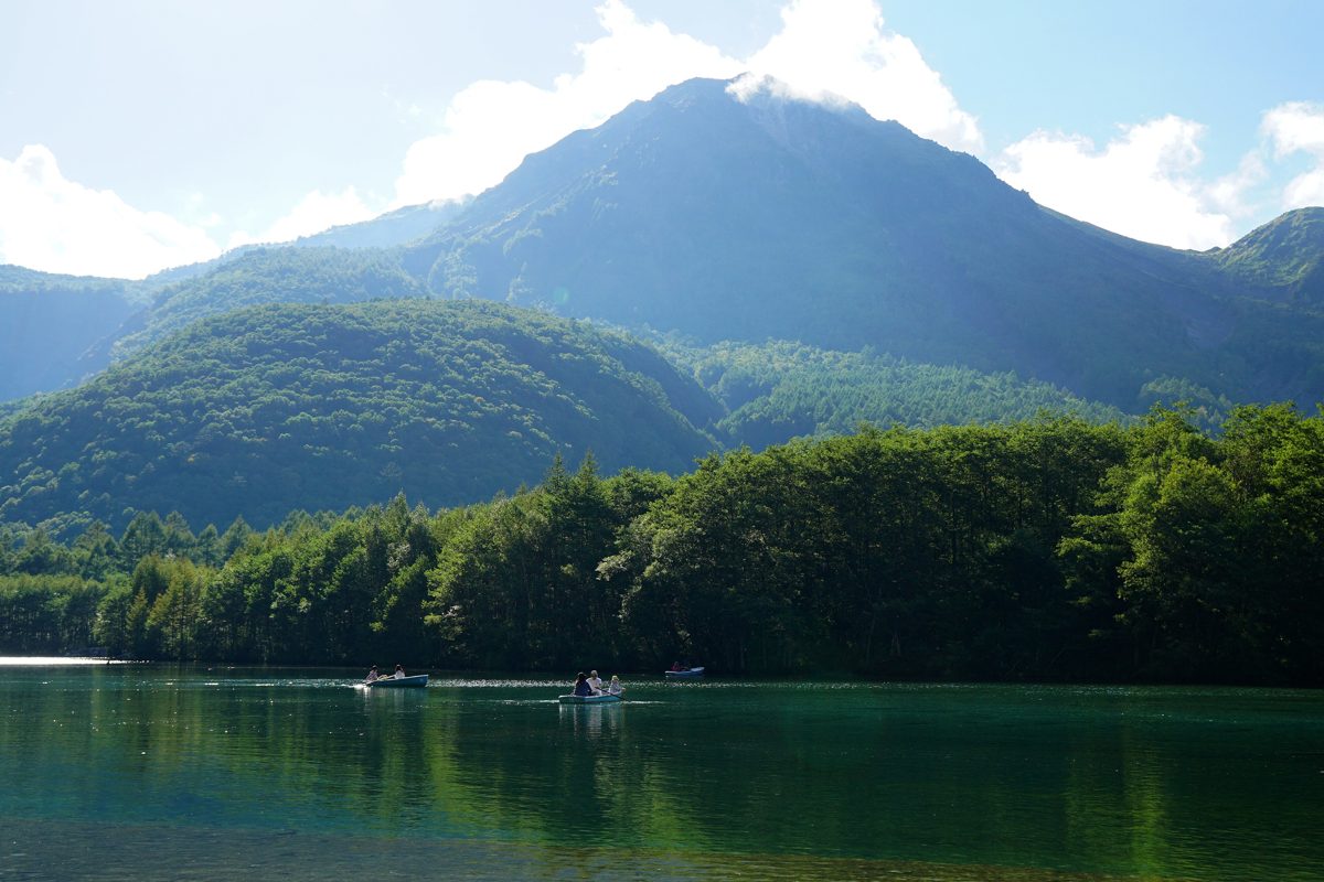 A calm green pond with a forested mountain behind it under a blue sky