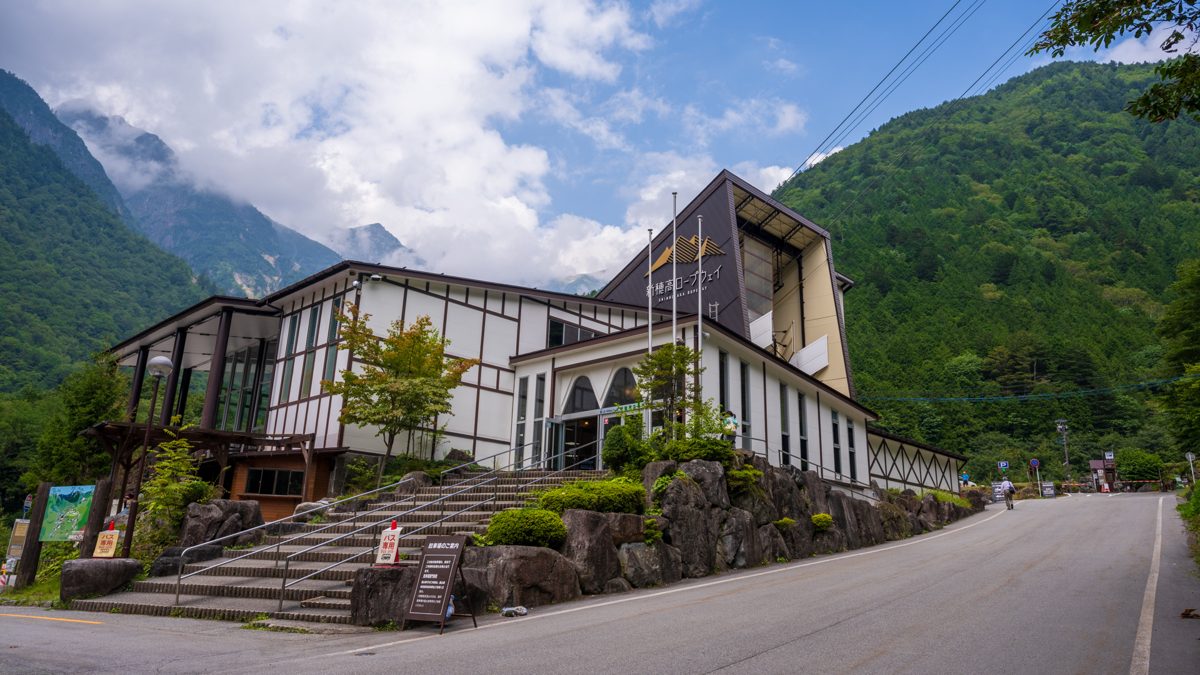 Shin-Hotaka Onsen Station of the Shinhotaka Ropeway, a wood-trimmed building beside a road below green mountains on a sunny day