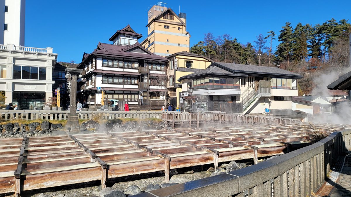 Wooden channels of steaming hot spring water in front of ryokan buildings under a blue sky