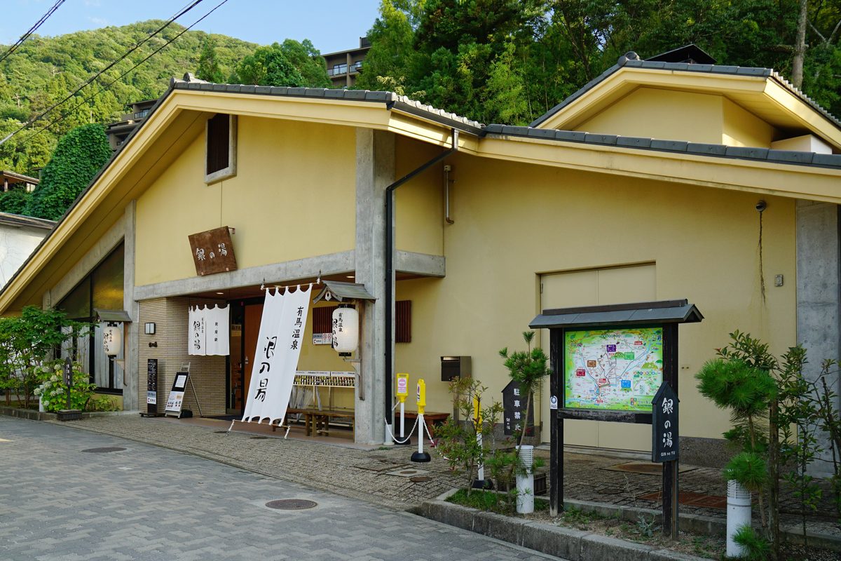 A cream-colored bathhouse with a white noren banner and a town map board