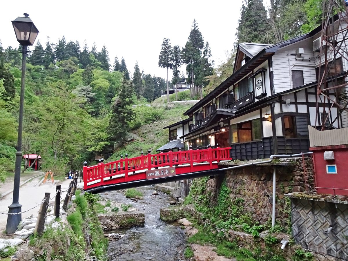 A red bridge over a stream between wooden ryokan buildings in a green valley