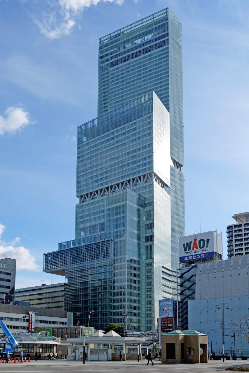 The tall stepped glass tower of Abeno Harukas against a blue sky above a station plaza