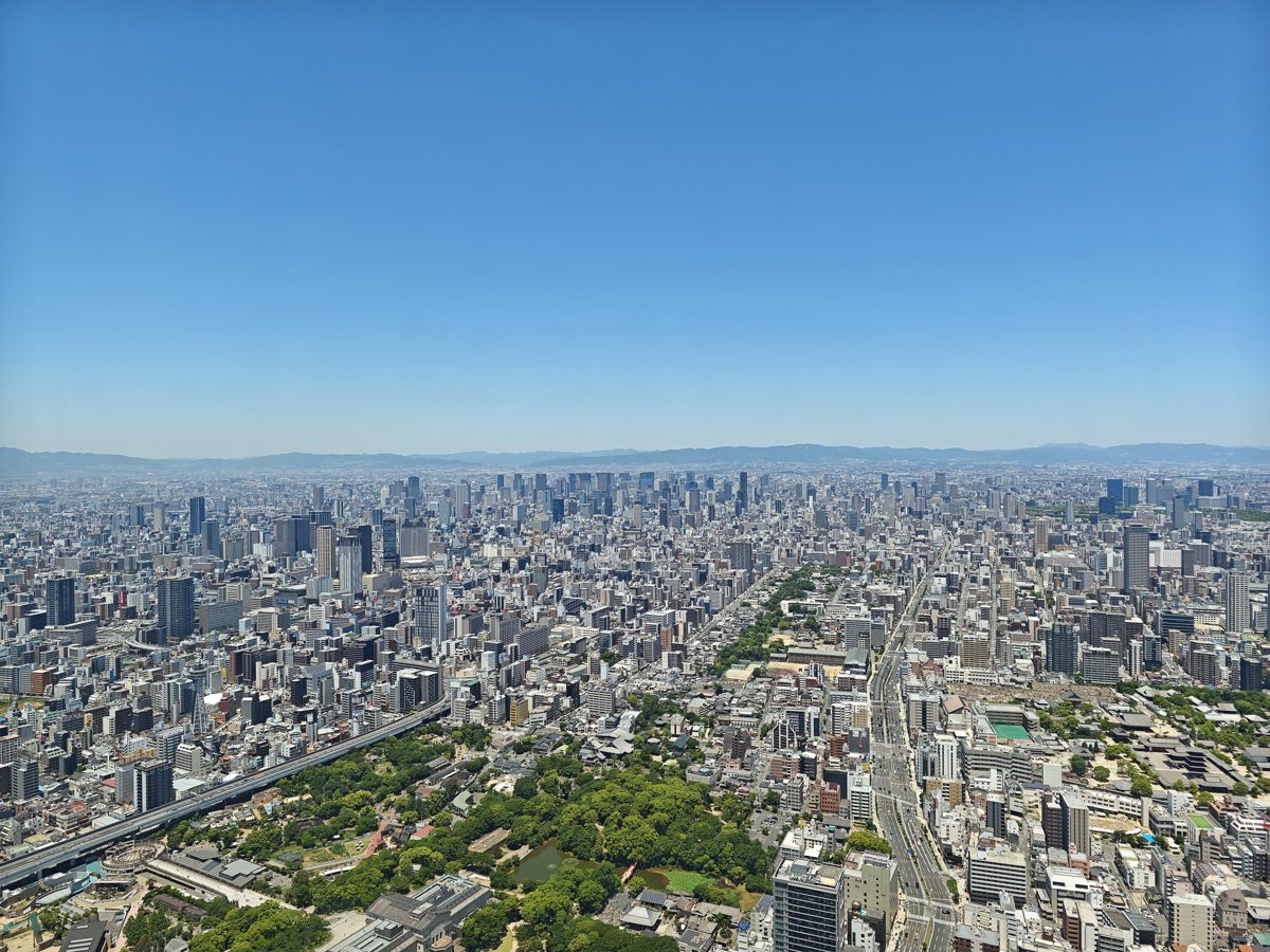 A wide view of central Osaka’s buildings and green park strips under a clear blue sky, seen from high above