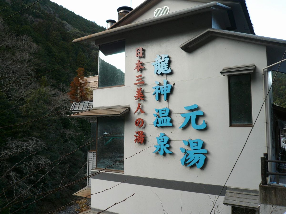 A white building with a large blue sign reading Ryujin Onsen Motoyu, against wooded hills