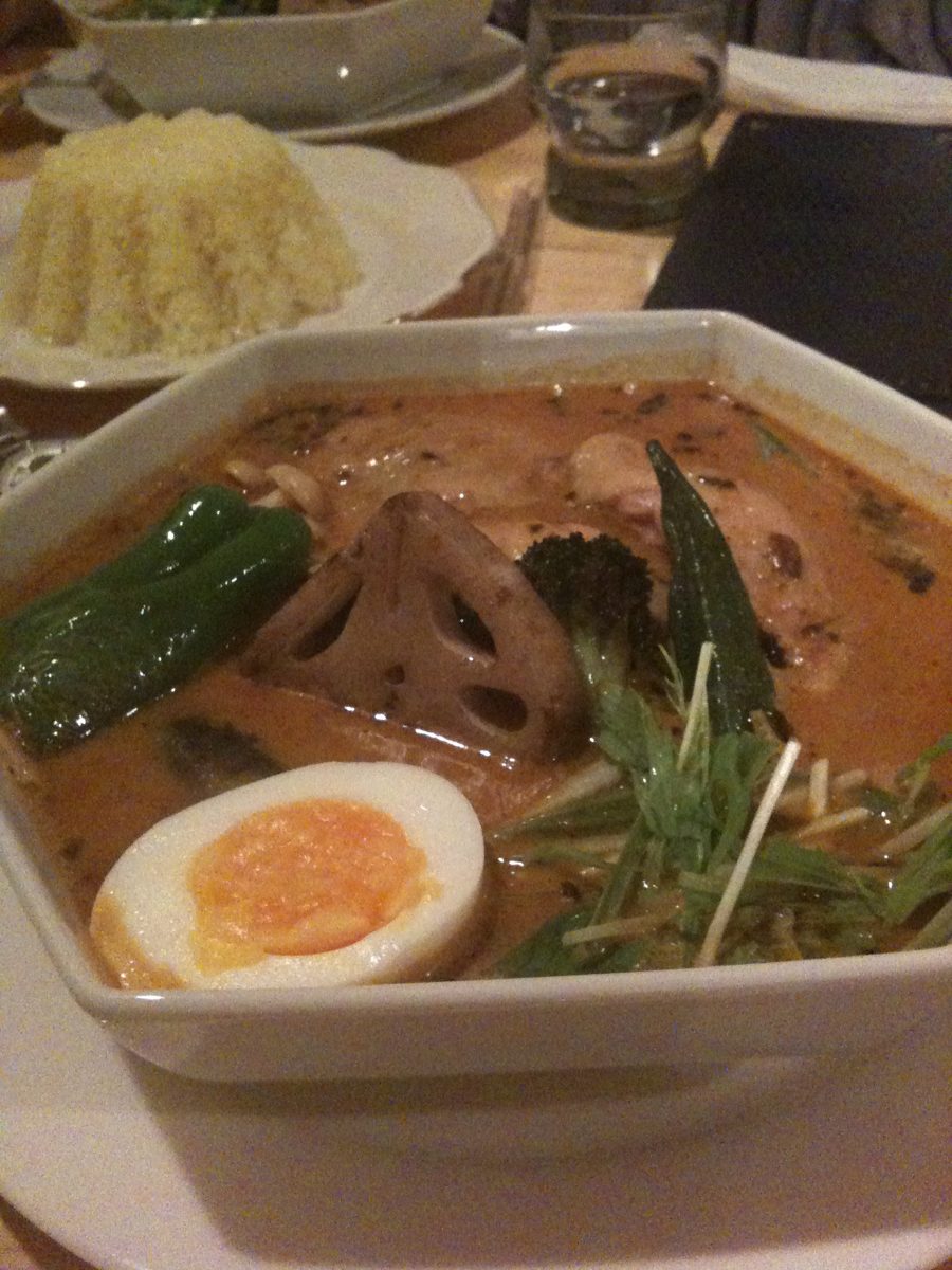 A bowl of soup curry with vegetables and a boiled egg, with rice behind