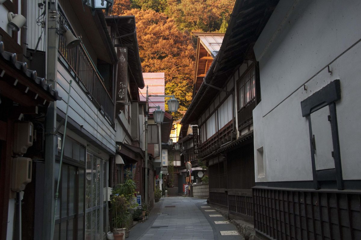 A narrow stone-paved lane between old wooden buildings in Shibu Onsen, with autumn trees on the hill behind